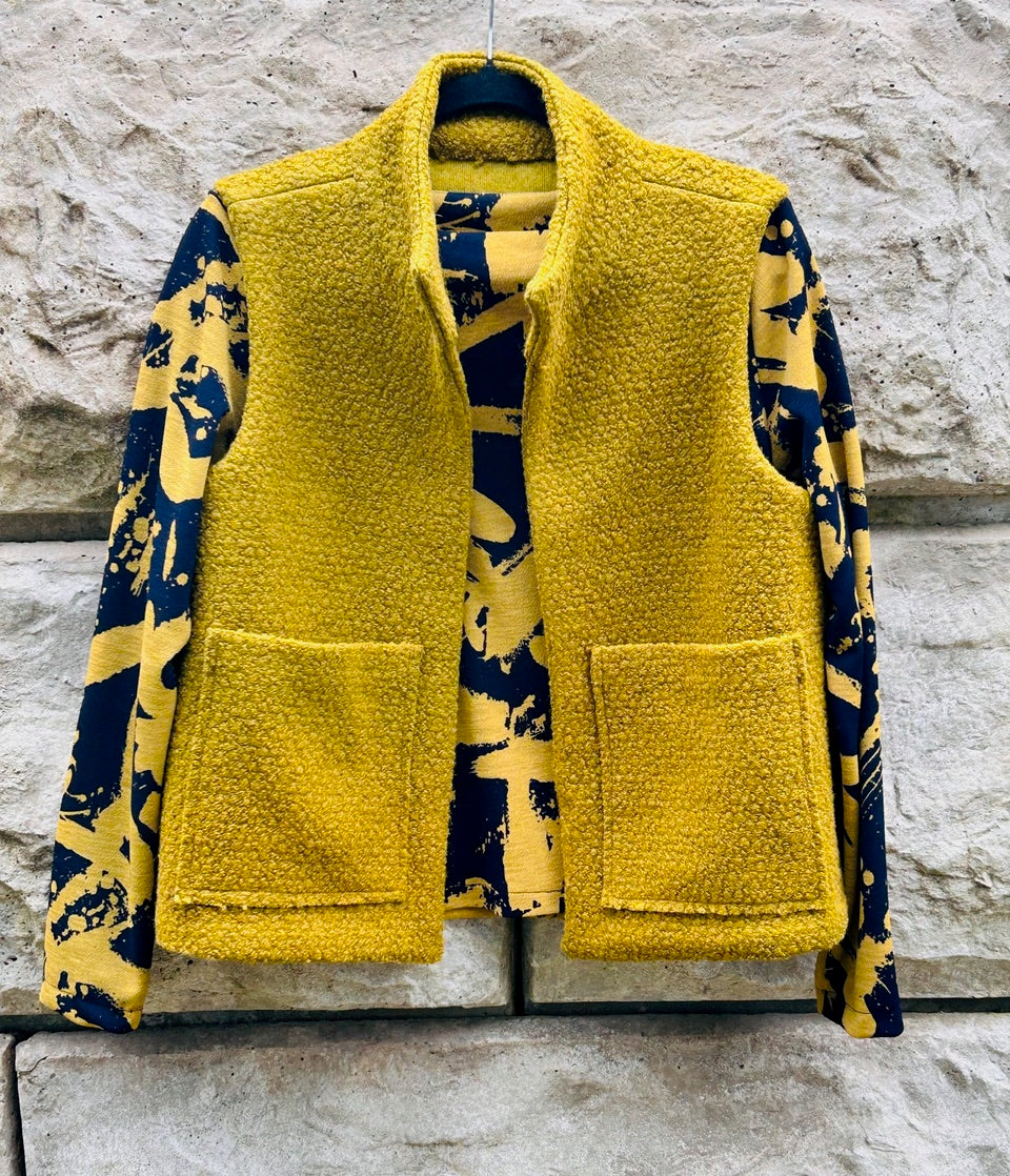 Yellow textured vest with front pockets over blue and yellow abstract print top, displayed on stone wall